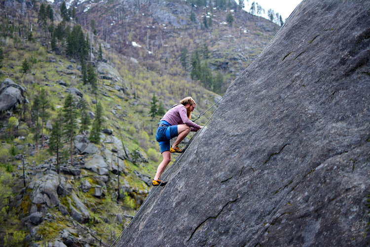 Eine Person klettert an einer steilen Felswand in einer grünen, bewaldeten Berglandschaft.