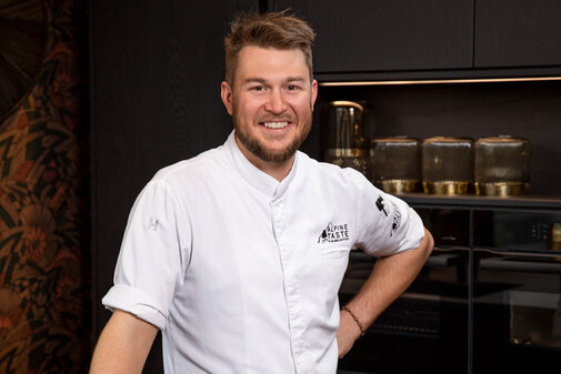 Ein lächelnder Koch in einer modernen Küche trägt eine weiße Kochjacke mit dem Logo 'Alpine Taste'.