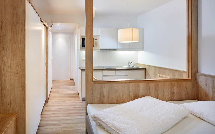 Der Raum zeigt ein gemütliches Bett mit weißen Kissen und eine offene Küche, die durch eine Holzverkleidung getrennt ist.