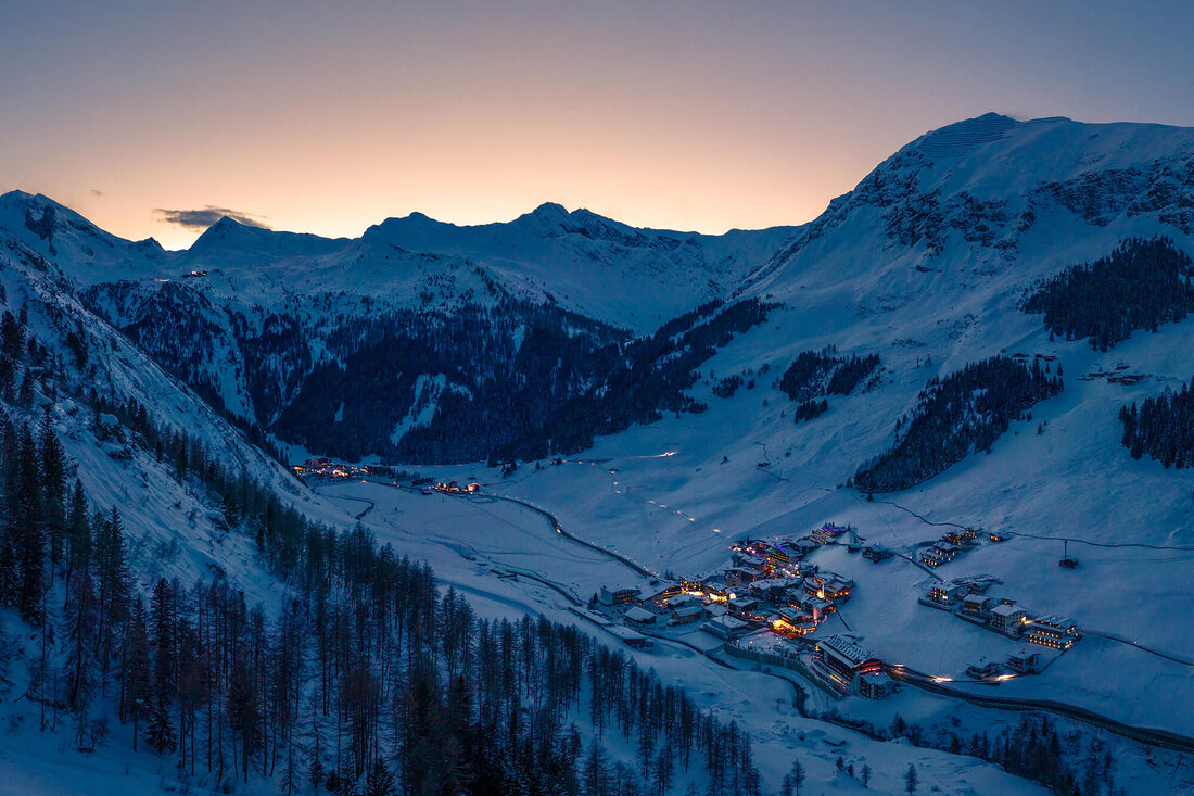 Die schneebedeckten Berge umgeben ein kleines, beleuchtetes Dorf in der Dämmerung, das eine friedliche Winteratmosphäre ausstrahlt.