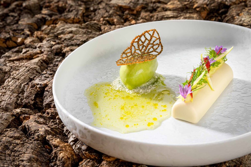 Ein elegantes Dessert auf einem weißen Teller, bestehend aus grünem Sorbet, einer knusprigen Blattdekoration und essbaren Blumen.