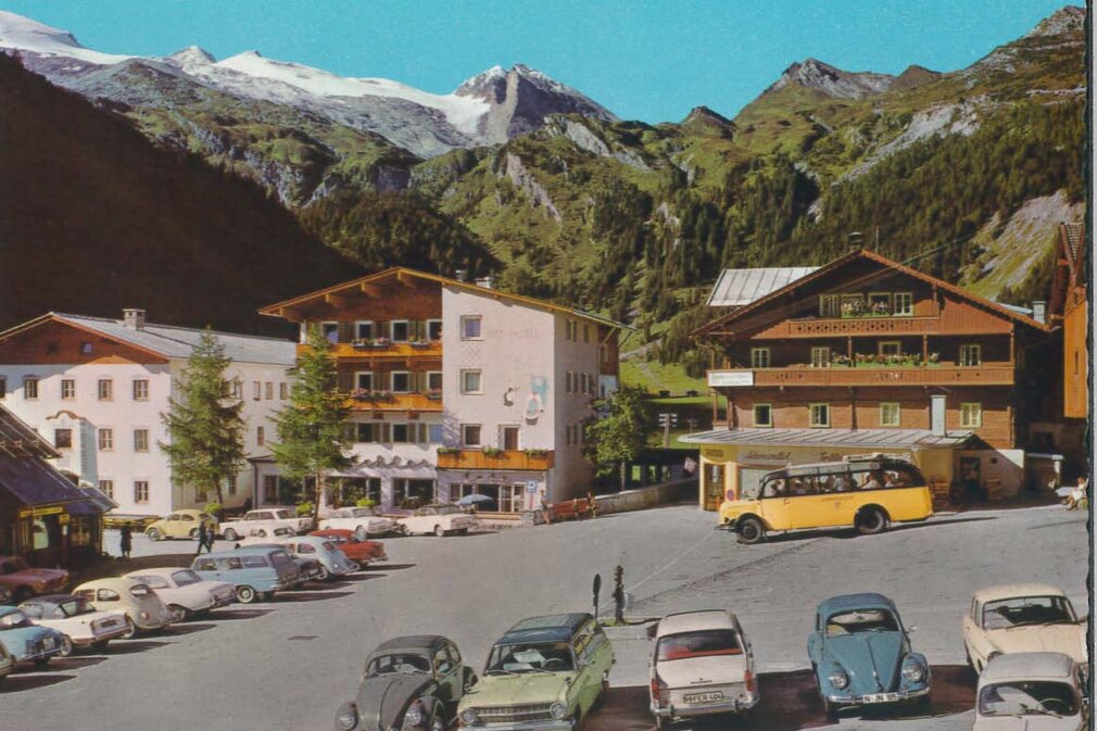 Autos parken auf einem Platz vor charmanten Gebäuden, umgeben von grünen Bäumen und majestätischen Bergen.