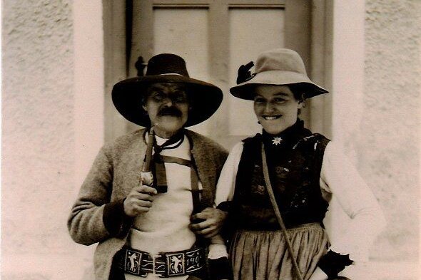 Ein lächelndes Paar in traditioneller Tracht steht vor einer Tür, der Mann trägt einen Hut und eine Weste, die Frau ein Kleid.