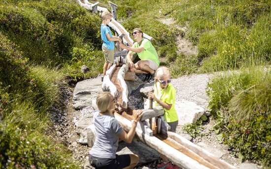 Eine Familie spielt im Freien, während Kinder an einem Wasserlauf experimentieren und ein Erwachsener ihnen hilft.