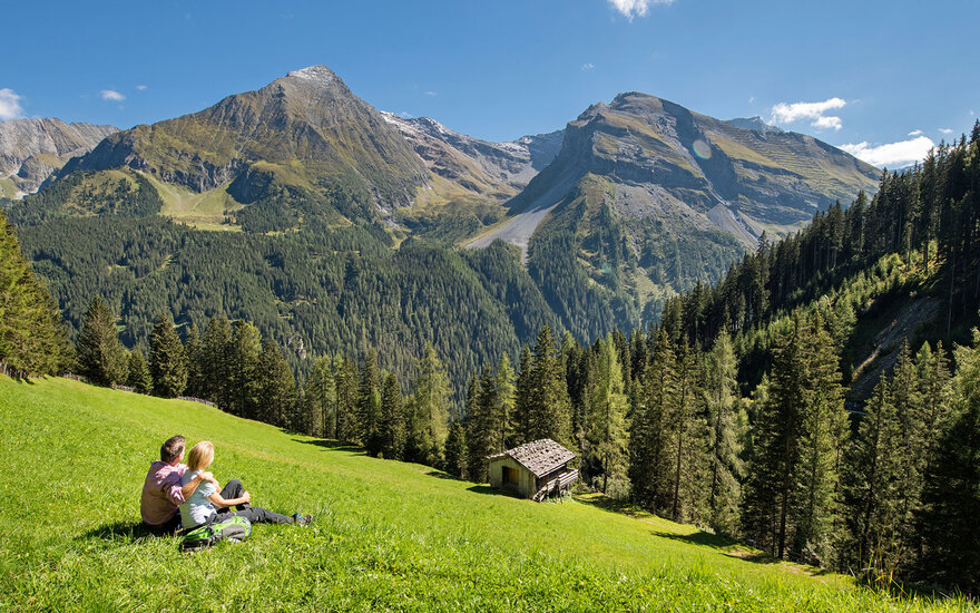 Ein Paar sitzt auf einer Wiese und genießt die Aussicht auf die majestätischen Berge und den klaren blauen Himmel.