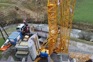 Auf der Baustelle sind ein gelber Kran und ein Lkw zu sehen, umgeben von Baumaterialien und einer malerischen Landschaft.