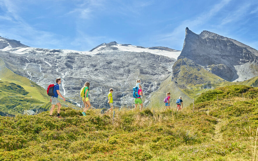 Eine Familie wandert fröhlich auf einem grünen Hügel, umgeben von majestätischen Bergen und klarem blauen Himmel.