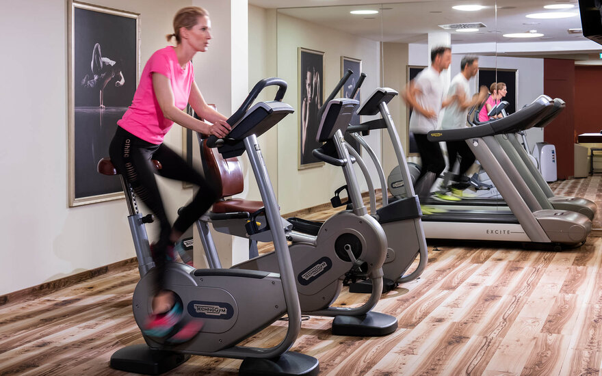 Eine Frau in einem pinken Shirt trainiert auf einem Hometrainer in einem modernen Fitnessraum mit Holzfußboden.