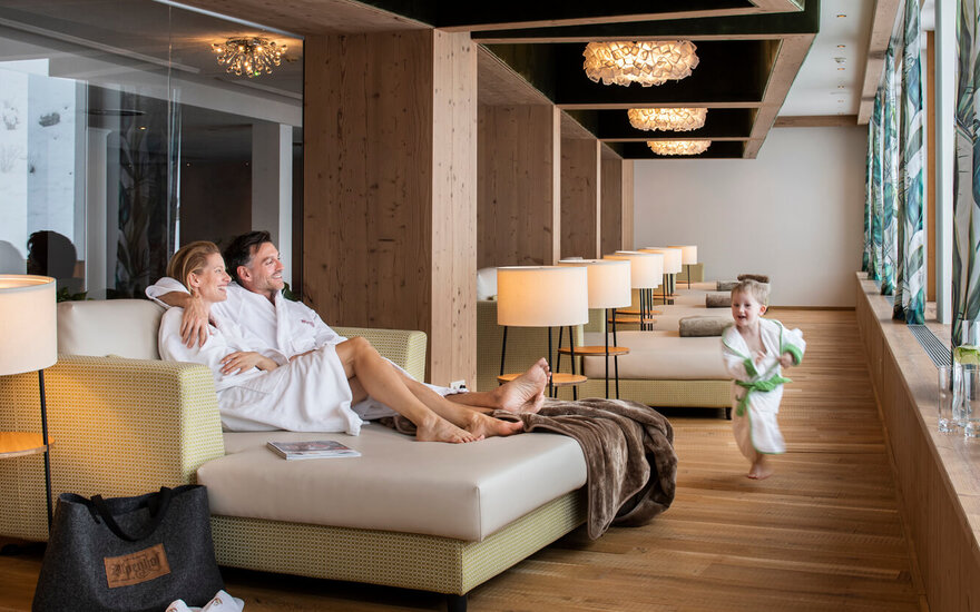 Das Bild zeigt eine gemütliche Lounge mit einem Paar, das sich entspannt, während ein Kind spielerisch vorbeiläuft.