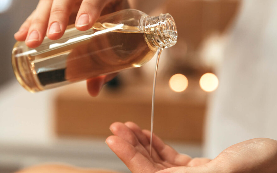 Eine Hand gießt ein goldenes Öl aus einer Flasche in die andere Hand, während im Hintergrund sanftes Licht leuchtet.