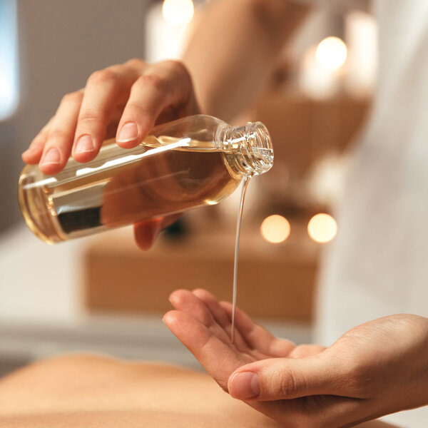 Eine Hand gießt ein goldenes Öl aus einer Flasche in die andere Hand, während im Hintergrund sanftes Licht leuchtet.
