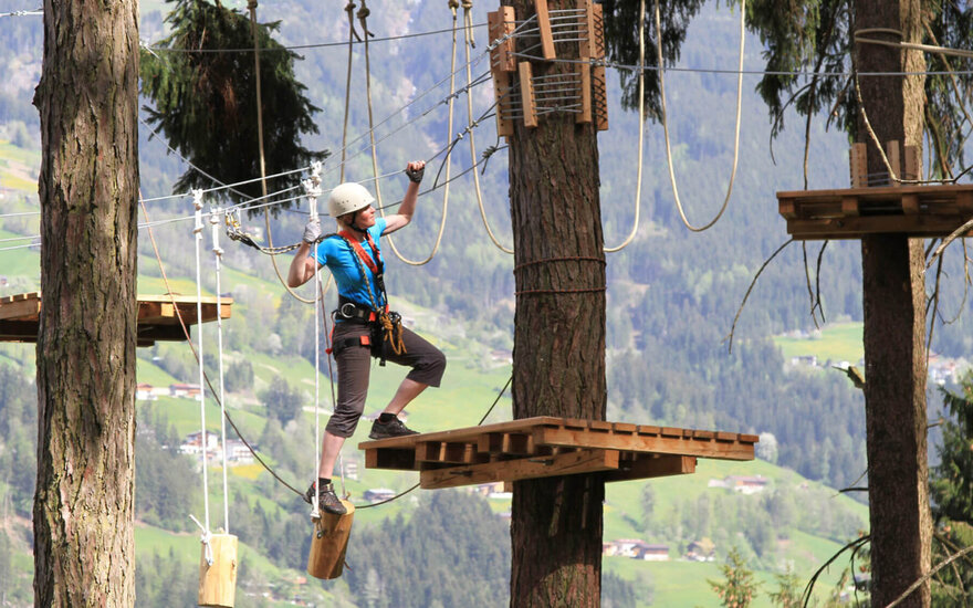 Eine Person in einem Kletterpark balanciert auf einem Baumstamm zwischen zwei Plattformen in der Natur.
