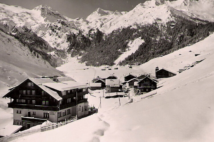 Schneebedeckte Hütten in einem ruhigen Tal, umgeben von hohen Bergen, die eine friedliche Winteratmosphäre schaffen.