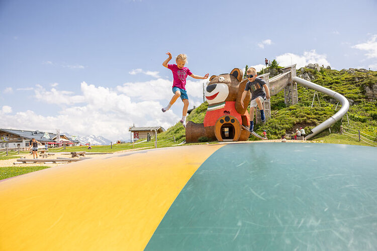 Zwei Kinder spielen auf einem bunten Trampolin, umgeben von einer malerischen Berglandschaft und einem Bärenhaus.