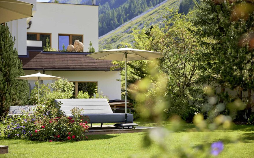 Ein elegantes Gartenmöbelstück mit einem Sonnenschirm steht in einem blühenden Garten, umgeben von grünen Bäumen und Bergen.