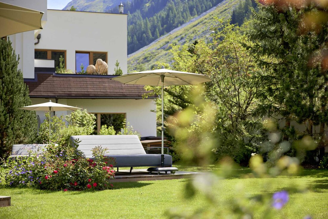 Ein elegantes Gartenmöbelstück mit einem Sonnenschirm steht in einem blühenden Garten, umgeben von grünen Bäumen und Bergen.