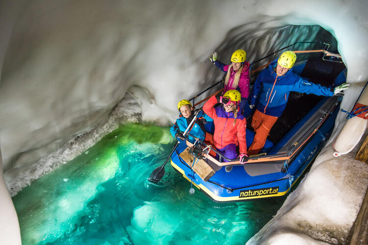 Eine Gruppe von vier Personen in bunten Anzügen posiert fröhlich in einem Boot in einer eisigen Höhle mit grünem Wasser.