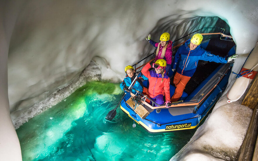 Eine Gruppe von vier Personen in bunten Anzügen posiert fröhlich in einem Boot in einer eisigen Höhle mit grünem Wasser.
