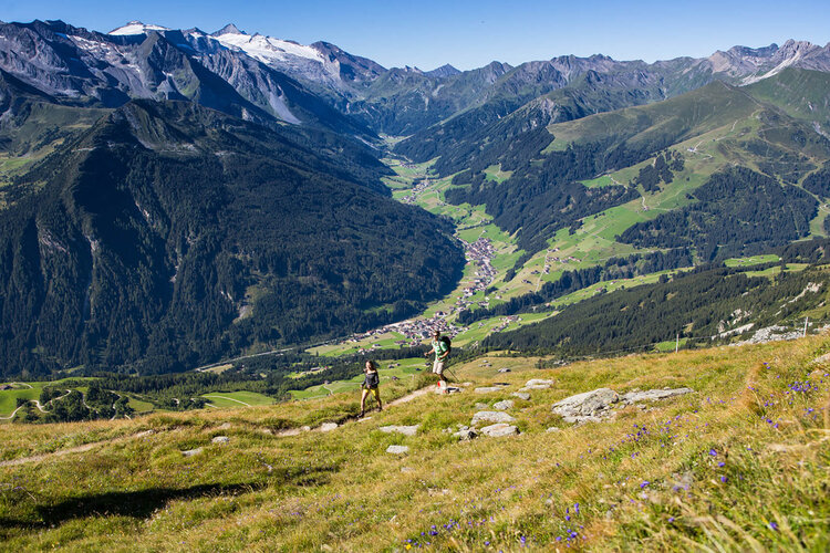 Zwei Wanderer erkunden die malerische Berglandschaft mit grünen Wiesen und schneebedeckten Gipfeln im Hintergrund.