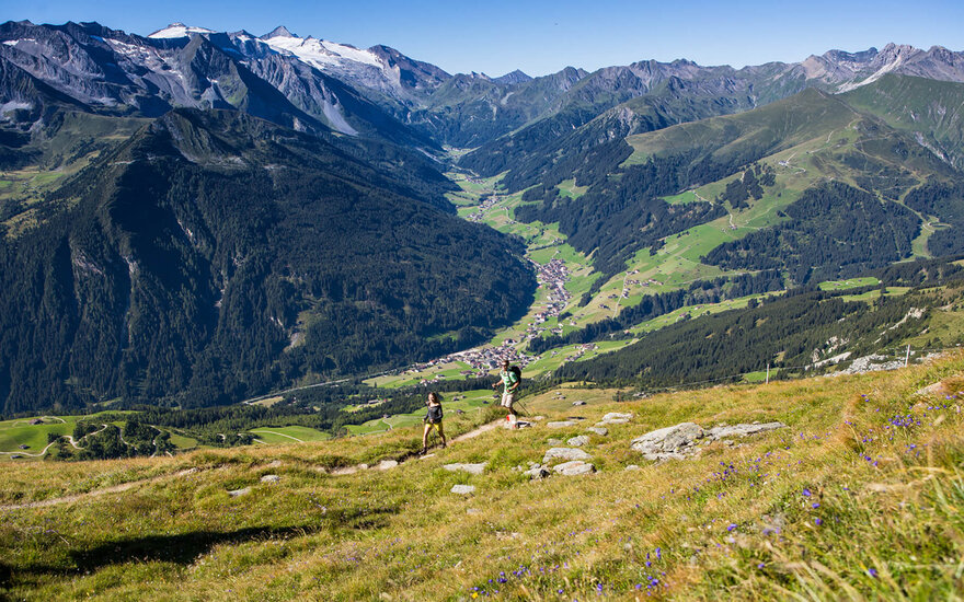 Zwei Wanderer erkunden die malerische Berglandschaft mit grünen Wiesen und schneebedeckten Gipfeln im Hintergrund.