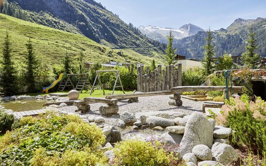Ein Spielplatz in den Alpen mit einer Rutsche, Schaukeln und einem Holzbalancierbalken, umgeben von grünen Wiesen und Bergen.