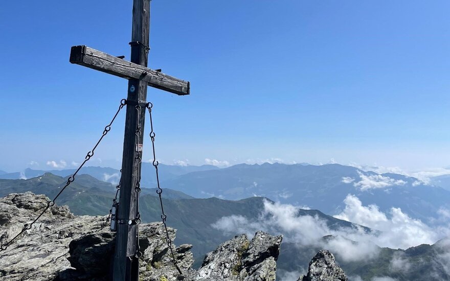 Das Kreuz, gesichert mit Ketten, thront über einer atemberaubenden Berglandschaft, die in der Ferne sanft verschwimmt.