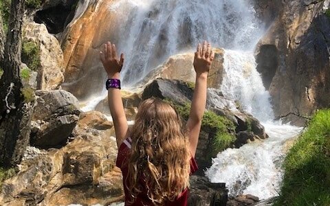 Ein Kind mit langen, lockigen Haaren steht vor einem beeindruckenden Wasserfall und hebt die Hände in die Luft.