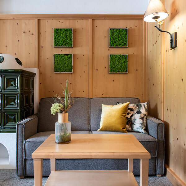 Ein gemütliches Wohnzimmer mit einem grauen Sofa, einem Holztisch und einer grünen Kachelofenheizung im Hintergrund.