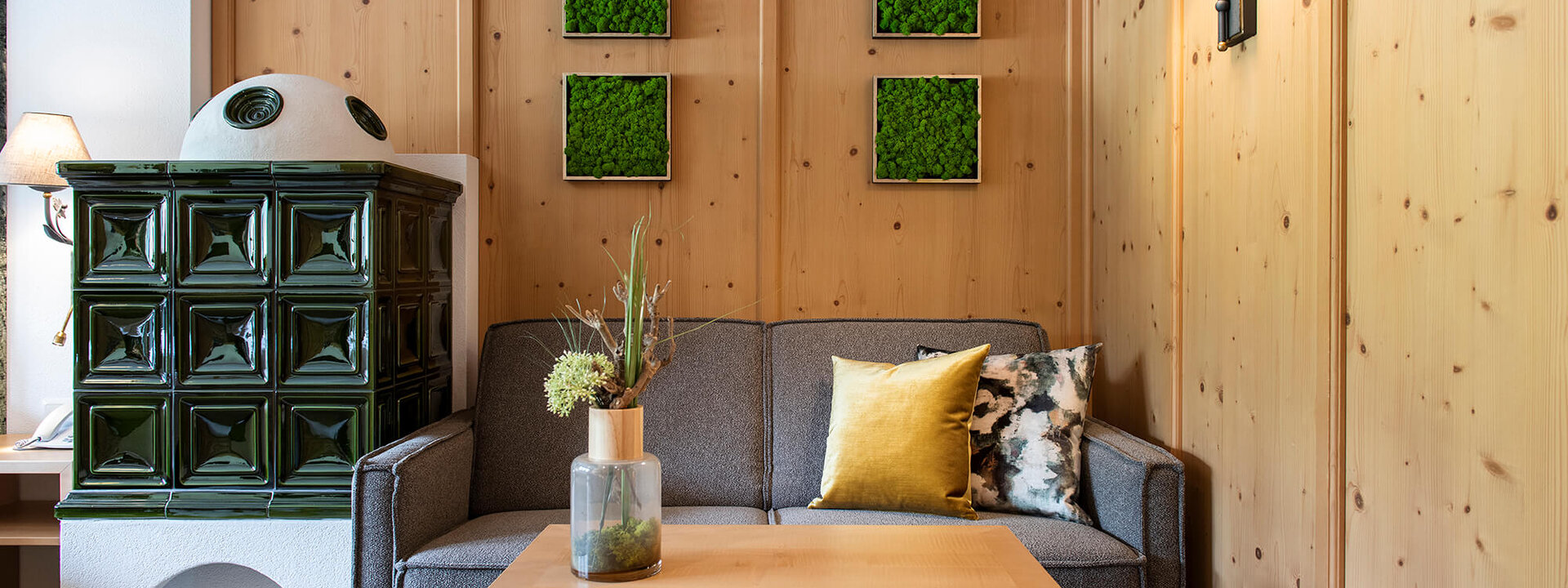 Ein gemütliches Wohnzimmer mit einem grauen Sofa, einem Holztisch und einer grünen Kachelofenheizung im Hintergrund.
