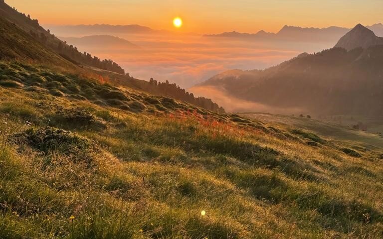 Ein atemberaubender Sonnenaufgang über sanften Hügeln, umgeben von Nebel und majestätischen Bergen im Hintergrund.