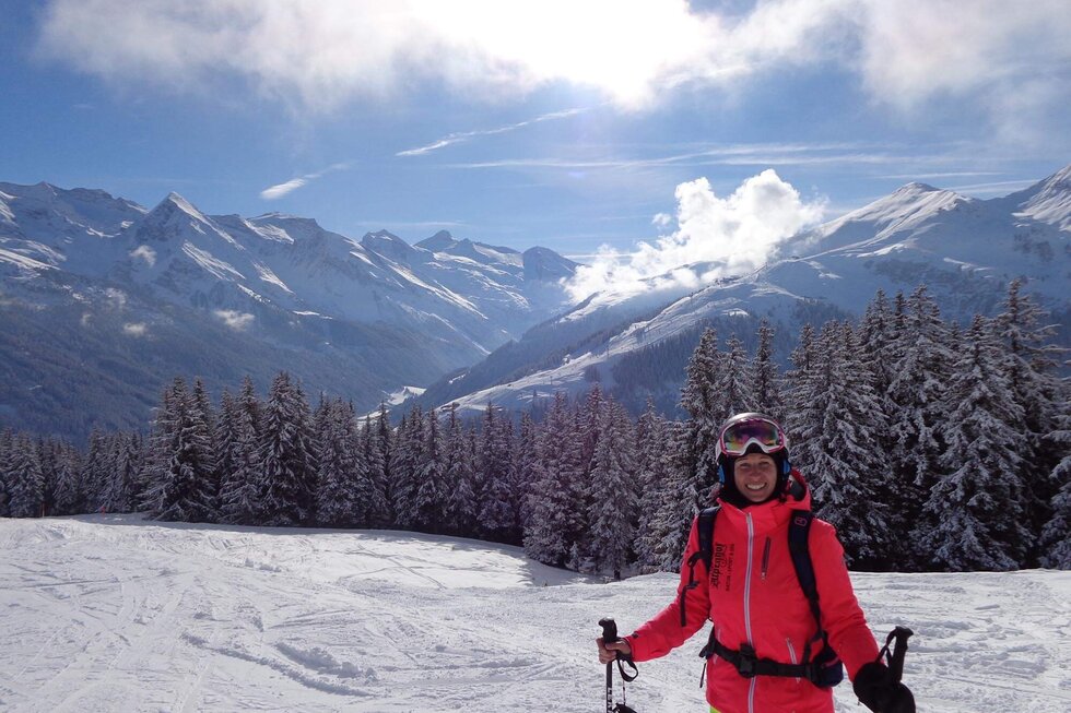 Ein fröhlicher Skifahrer in leuchtend pinker Jacke steht auf einer schneebedeckten Piste mit majestätischen Bergen im Hintergrund.