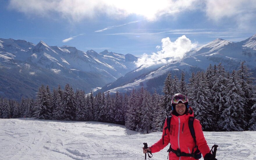 Ein fröhlicher Skifahrer in leuchtend pinker Jacke steht auf einer schneebedeckten Piste mit majestätischen Bergen im Hintergrund.
