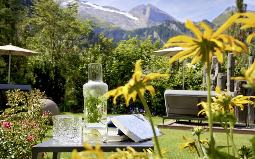 Ein Tisch im Garten mit einer Karaffe Wasser, Gläsern und einem Buch, umgeben von bunten Blumen und Bergen im Hintergrund.