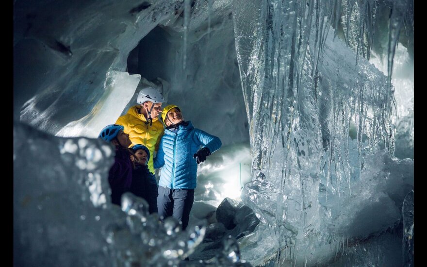 Eine Gruppe von vier Personen steht in einer Eishöhle, umgeben von glitzernden Eisformationen und beleuchtet von sanftem Licht.