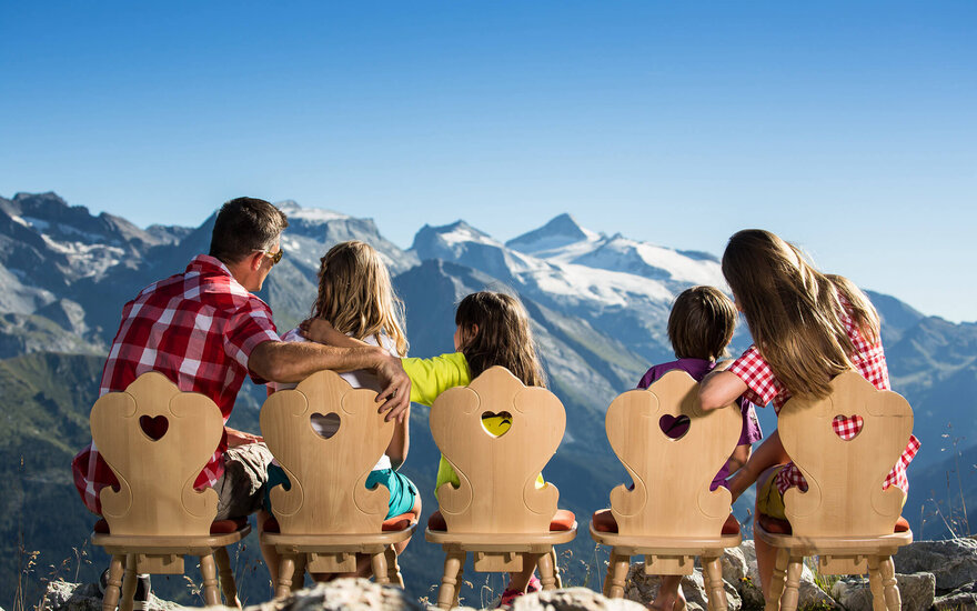 Eine Familie sitzt auf Holzstühlen mit Herzmotiven und blickt auf majestätische Berge unter klarem Himmel.