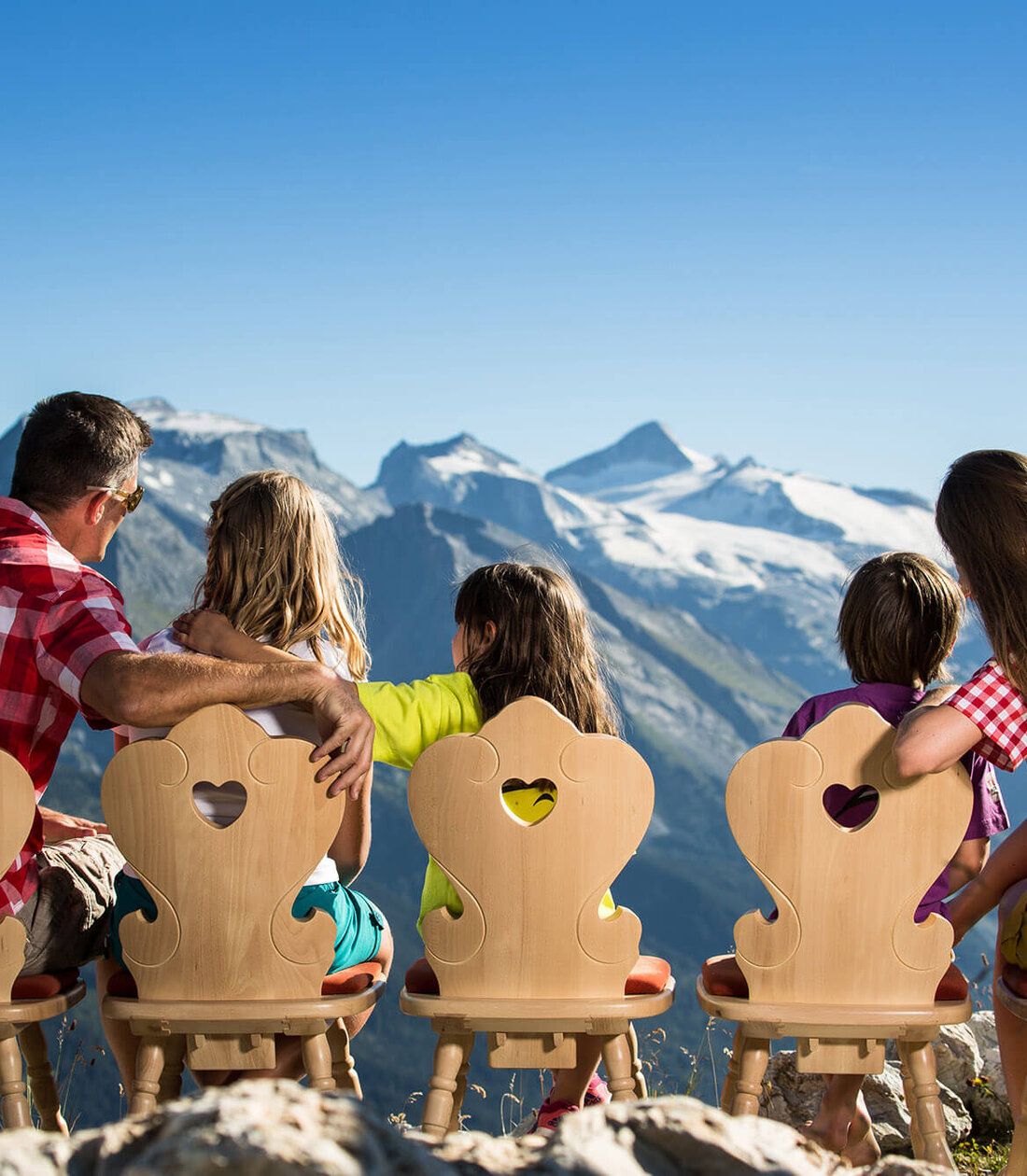 Eine Familie sitzt auf Holzstühlen mit Herzmotiven und blickt auf majestätische Berge unter klarem Himmel.