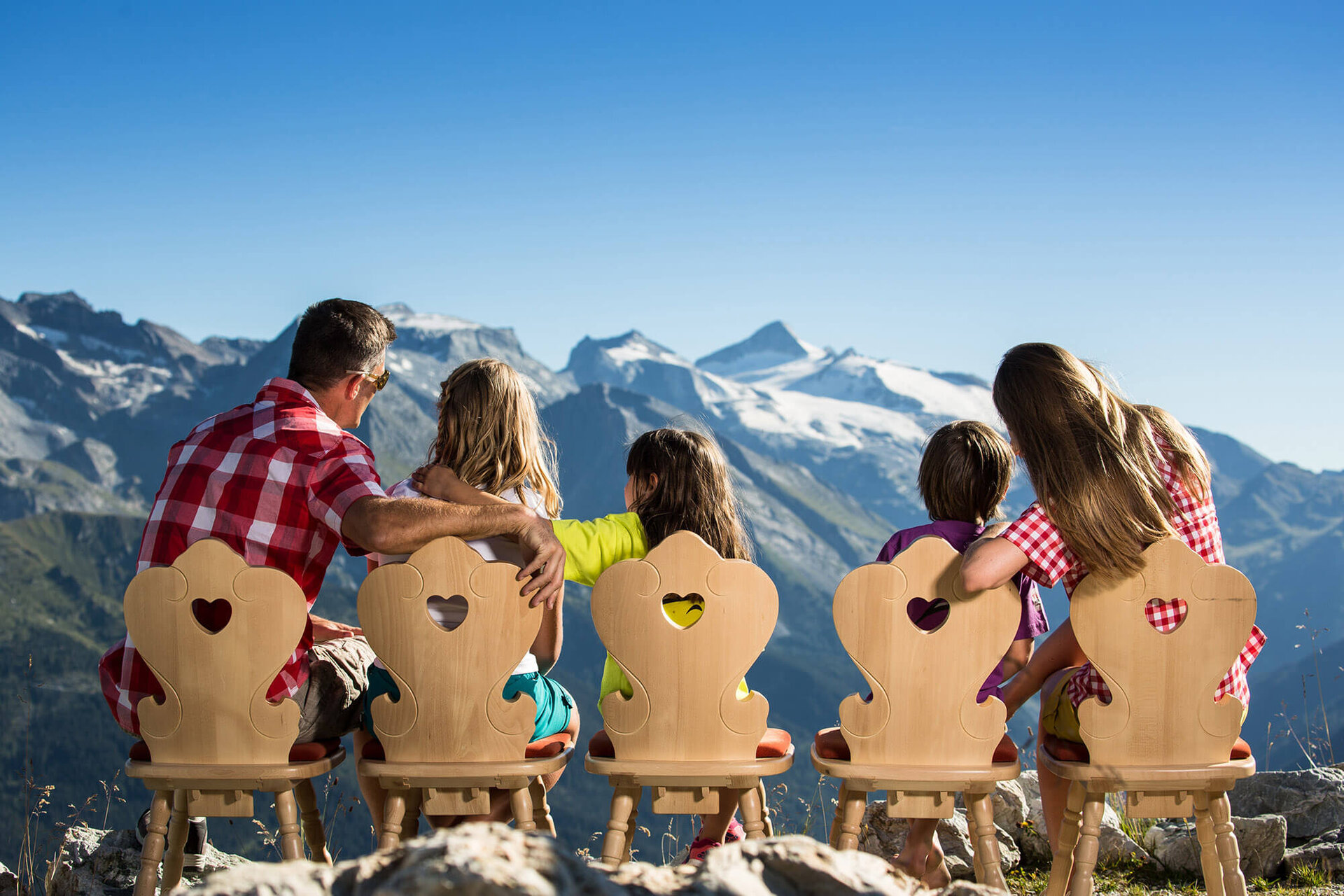 Eine Familie sitzt auf Holzstühlen mit Herzmotiven und blickt auf majestätische Berge unter klarem Himmel.