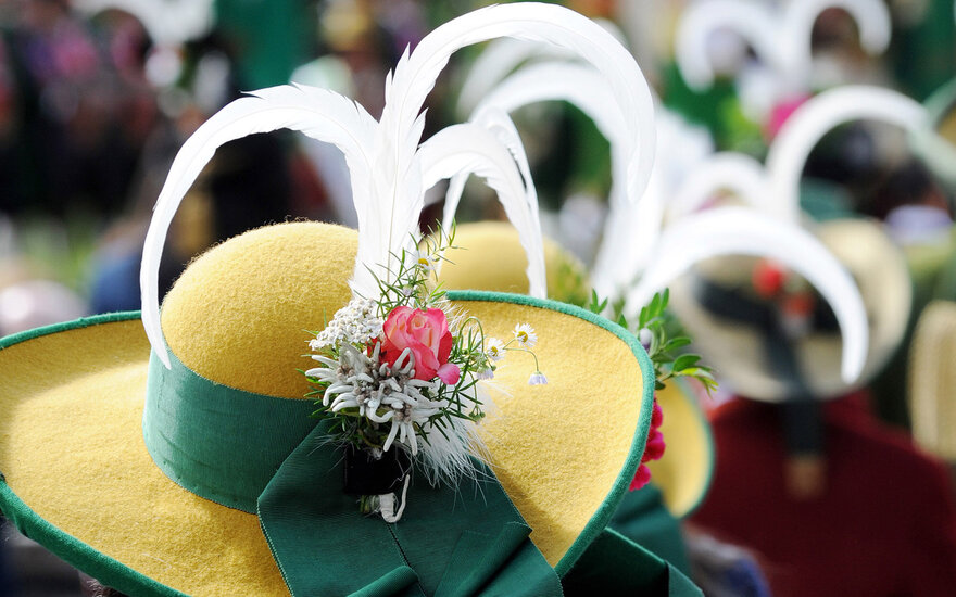 Ein traditioneller Hut mit gelber Krempe und üppiger Blumenverzierung, umgeben von weiteren Hüten in einer festlichen Atmosphäre.