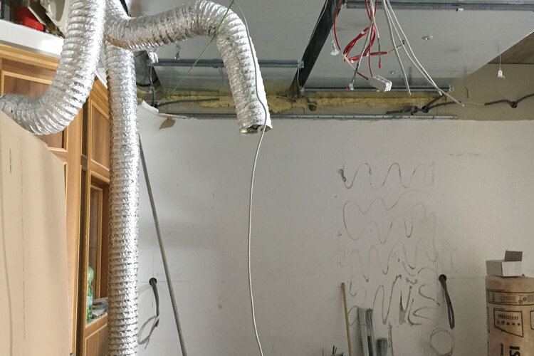Ein Raum mit einer silbernen Lüftungsleitung, die von der Decke hängt, und verschiedenen Werkzeugen und Materialien auf dem Boden.