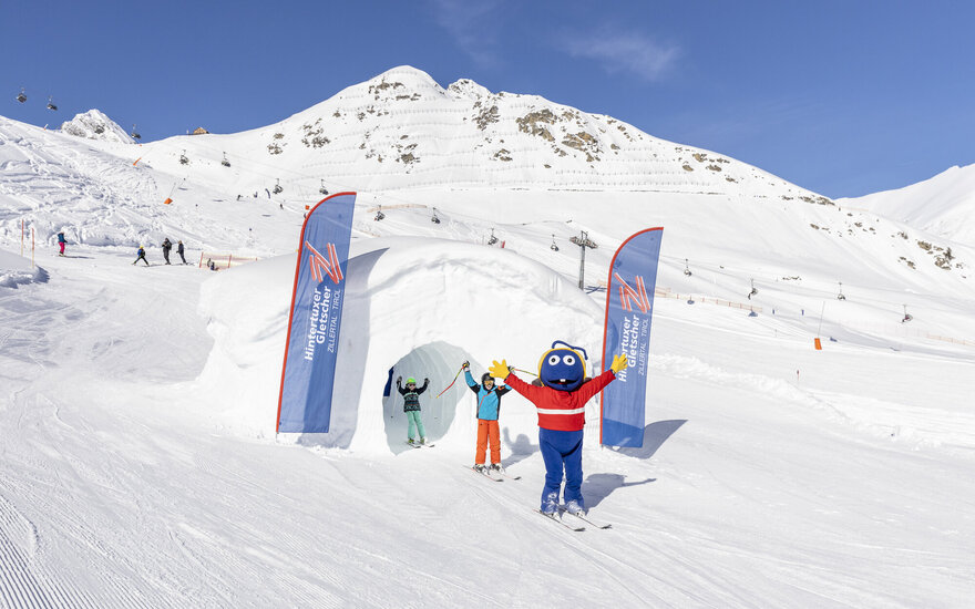 Ein fröhlicher Skimaskottchen begrüßt Kinder auf der Piste, umgeben von schneebedeckten Bergen und blauen Himmel.