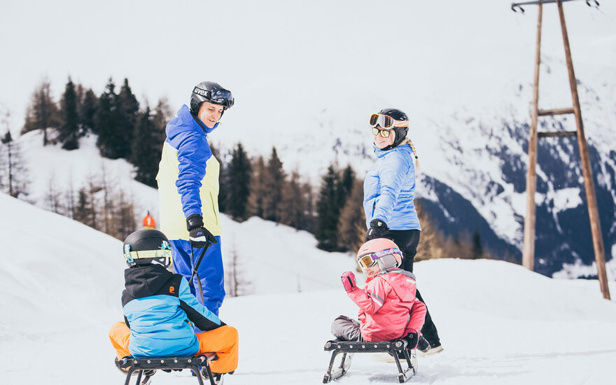 Eine Familie genießt einen sonnigen Tag im Schnee, während die Kinder auf Schlitten sitzen und fröhlich lachen.