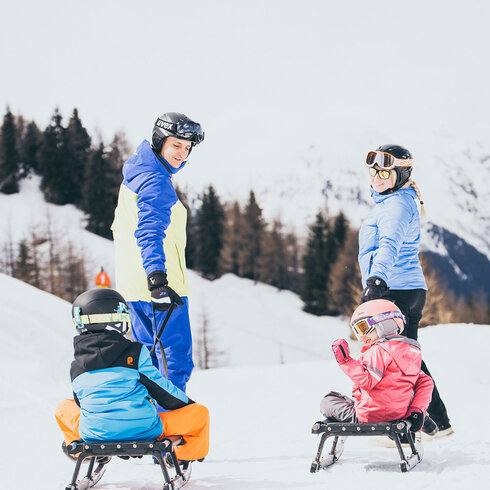 Eine Familie genießt einen sonnigen Tag im Schnee, während die Kinder auf Schlitten sitzen und fröhlich lachen.