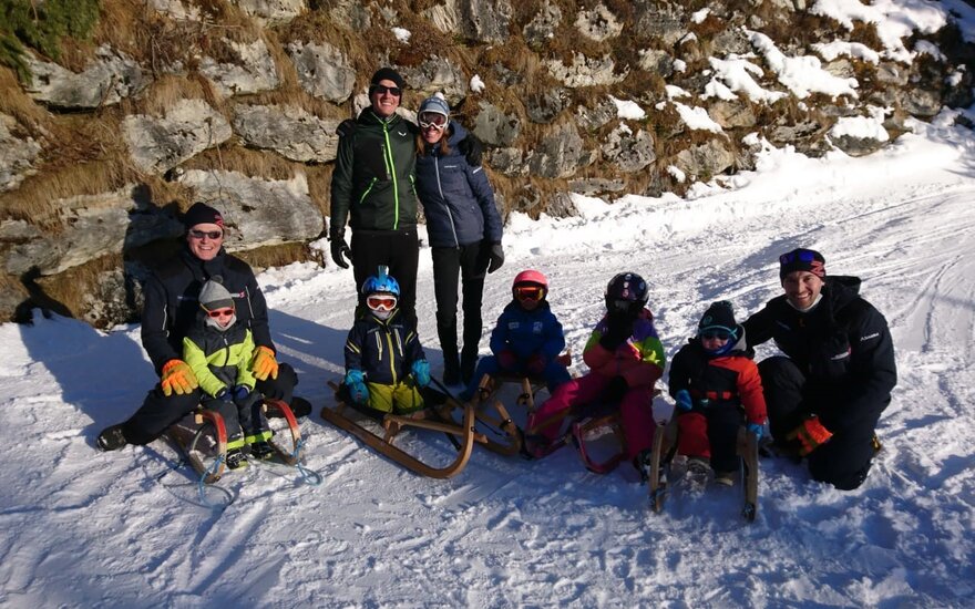 Eine fröhliche Familie mit Kindern sitzt auf Schlitten im Schnee, umgeben von einer felsigen Landschaft und strahlendem Sonnenschein.