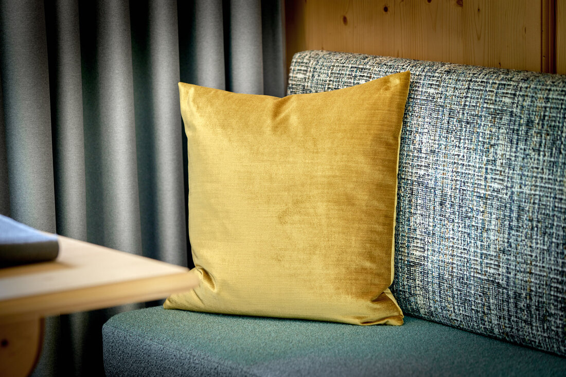 Ein leuchtend gelbes Kissen ruht auf einer blauen Couch, umgeben von grauen Vorhängen und einer Holzverkleidung.