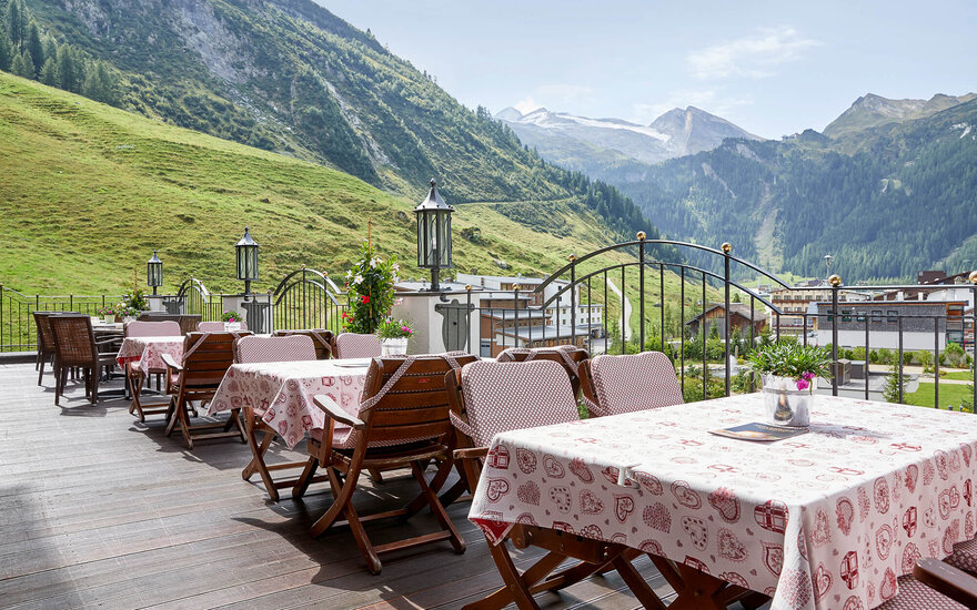 Eine sonnige Terrasse mit Holztischen und Stühlen, umgeben von grünen Hügeln und Bergen im Hintergrund.