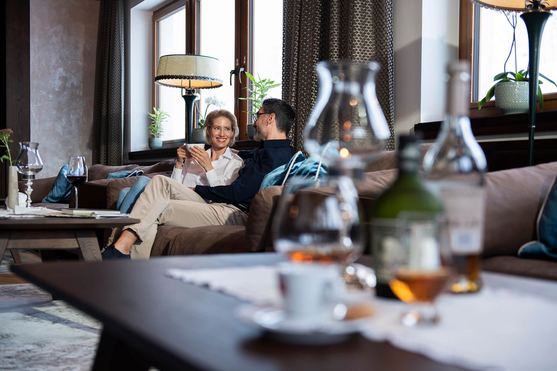 Ein Paar sitzt entspannt auf einem Sofa, während sie sich liebevoll anlächeln und eine Tasse in der Hand halten.