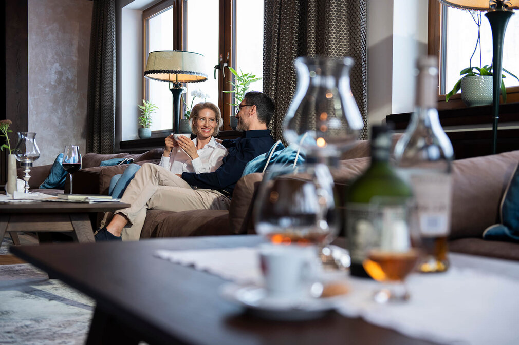 Ein Paar sitzt entspannt auf einem Sofa, während sie sich liebevoll anlächeln und eine Tasse in der Hand halten.
