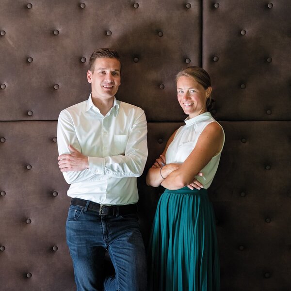 Vor einer stilvollen, braunen Wand stehen ein Mann und eine Frau, beide mit verschränkten Armen und freundlichen Gesichtsausdrücken.