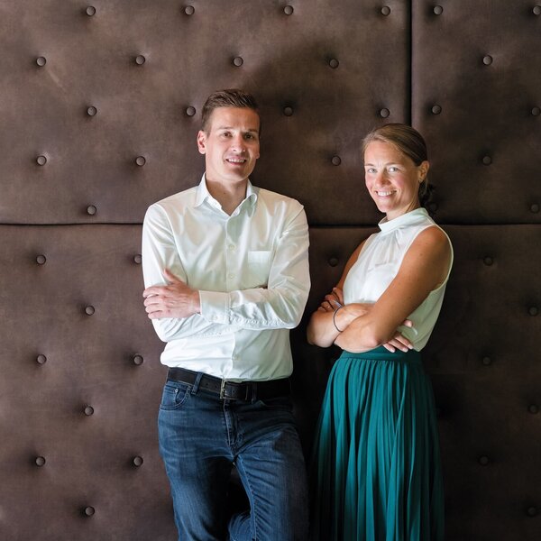 Vor einer stilvollen, braunen Wand stehen ein Mann und eine Frau, beide mit verschränkten Armen und freundlichen Gesichtsausdrücken.