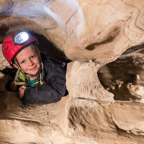 Ein Junge mit einem roten Helm lächelt, während er in einer engen Höhle sitzt und von einer Lampe beleuchtet wird.
